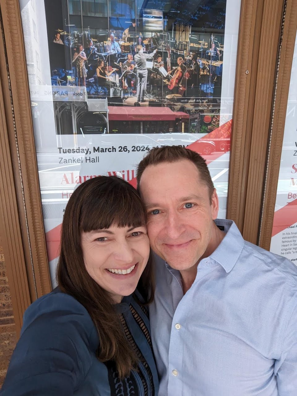 Erin Lesser and Michael Clayville take a selfie in front of an Alarm Will Sound concert poster.