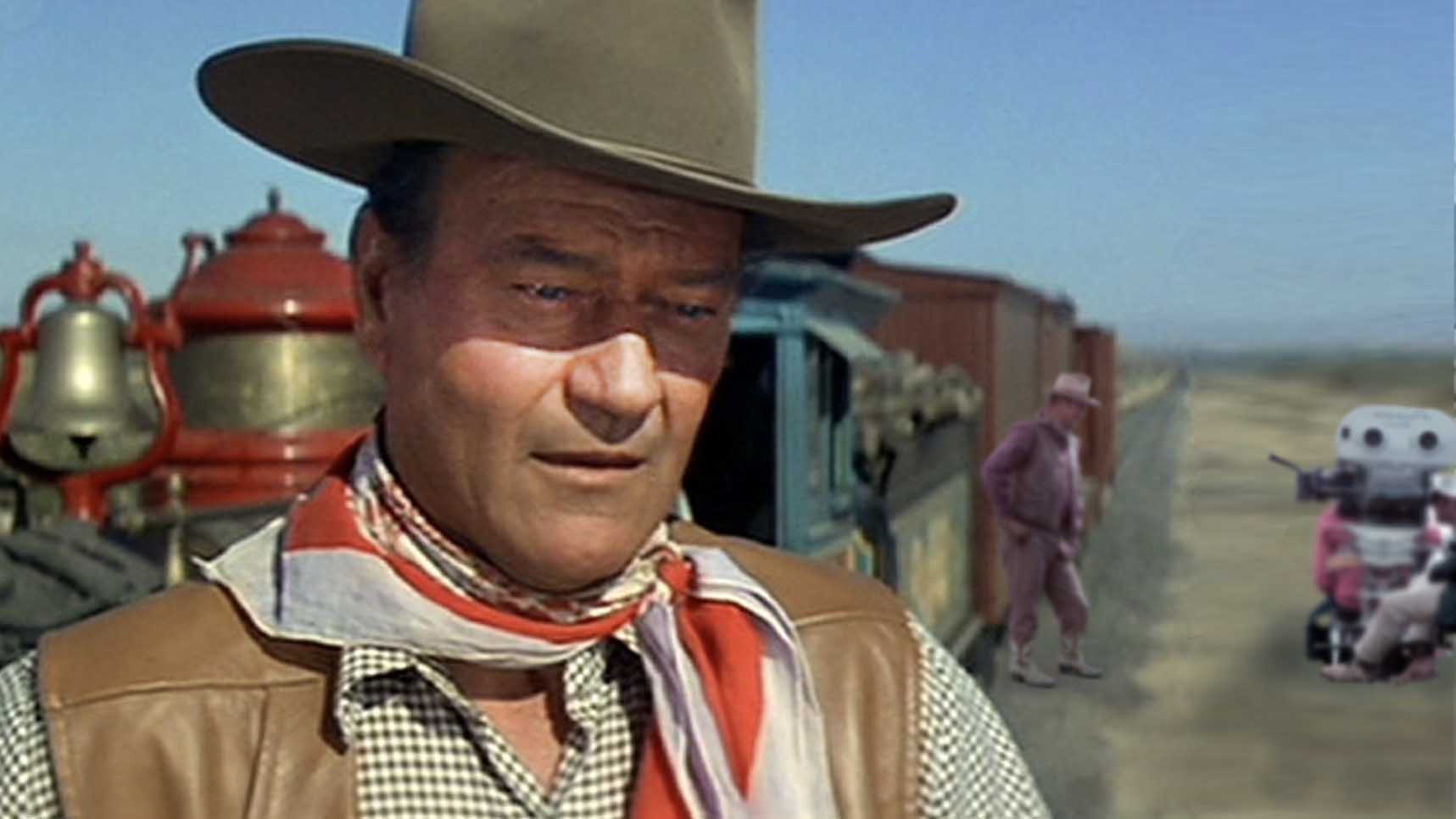 Actor John Wayne in foreground and and actor an camera crew in background in front of train