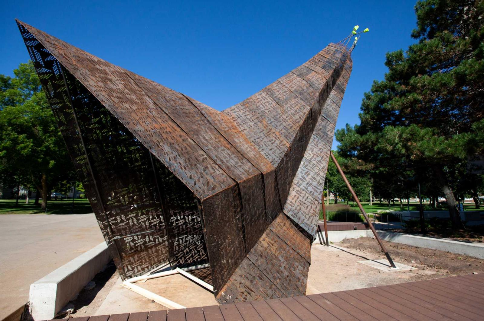 Giant sculpture with perforated and patinaed steel panels and geometric beadwork designs on them, overlap like a crane's feathers.