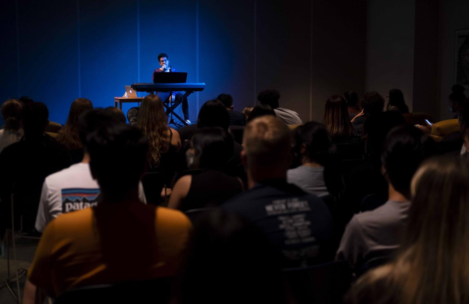 A student at an electronic keyboard under a blue spotlight sings on a makeshift stage while an audience watches.