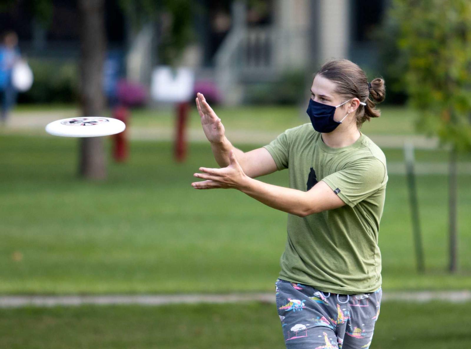 Student in mask reaching arms out to catch incoming frisbee on Main Hall Green.