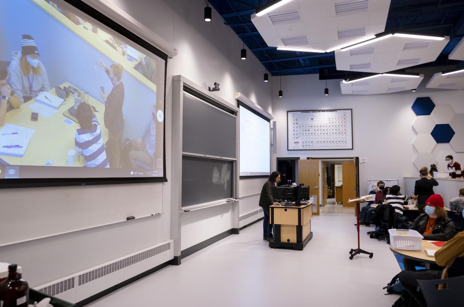 Physics professor lecturing in front of room as students sit in pods, working and taking notes.