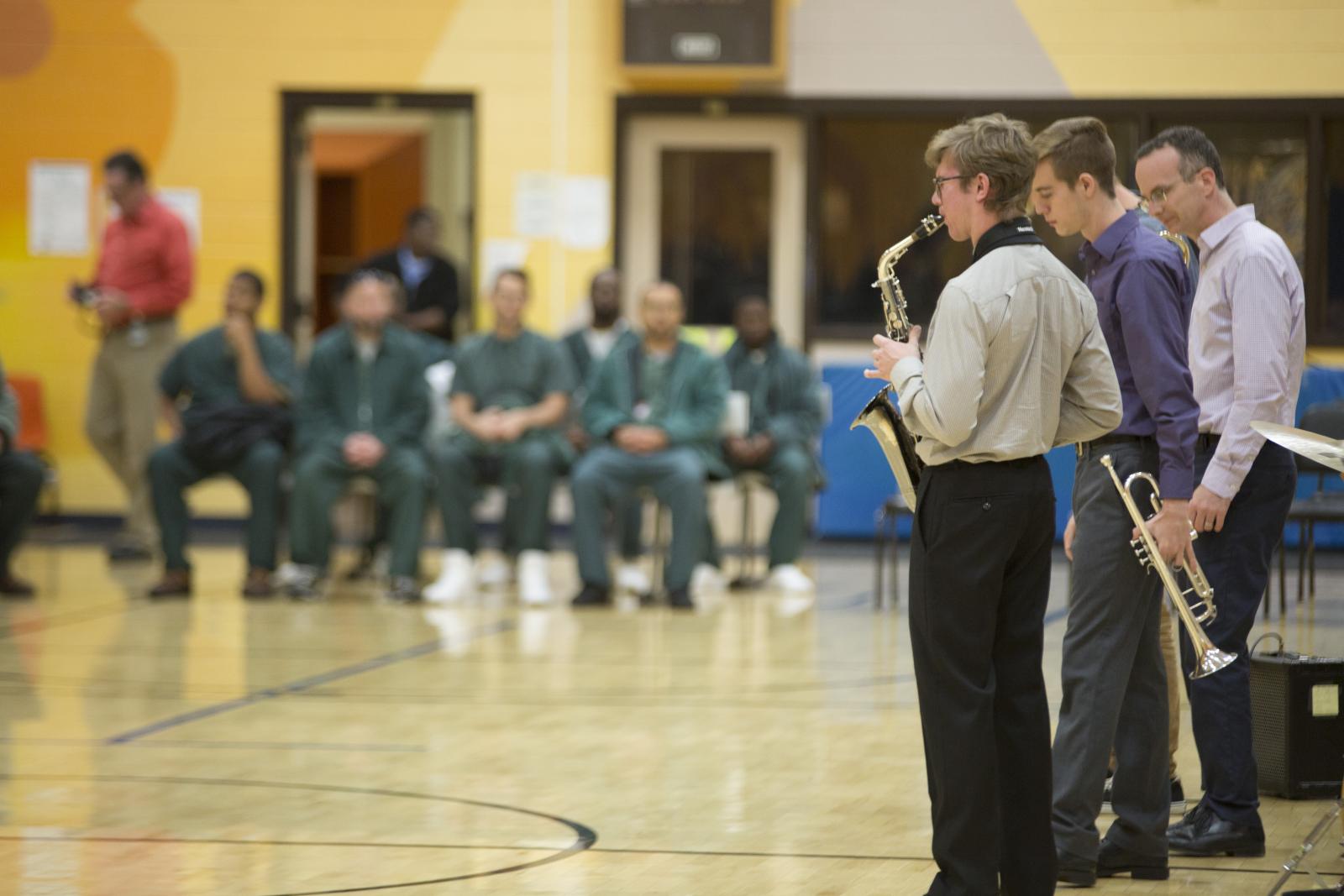 Jazz musicians play in a gynasium, with men in green jumpsuites in the background.