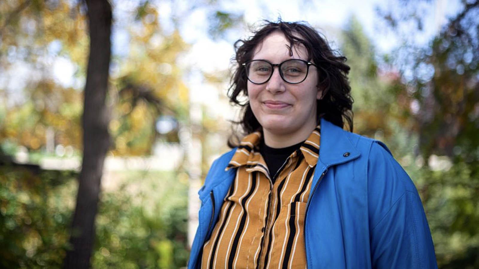 Dani Massey, wearing a blue jacket and yellow and black striped shirt, smiles at the camera.