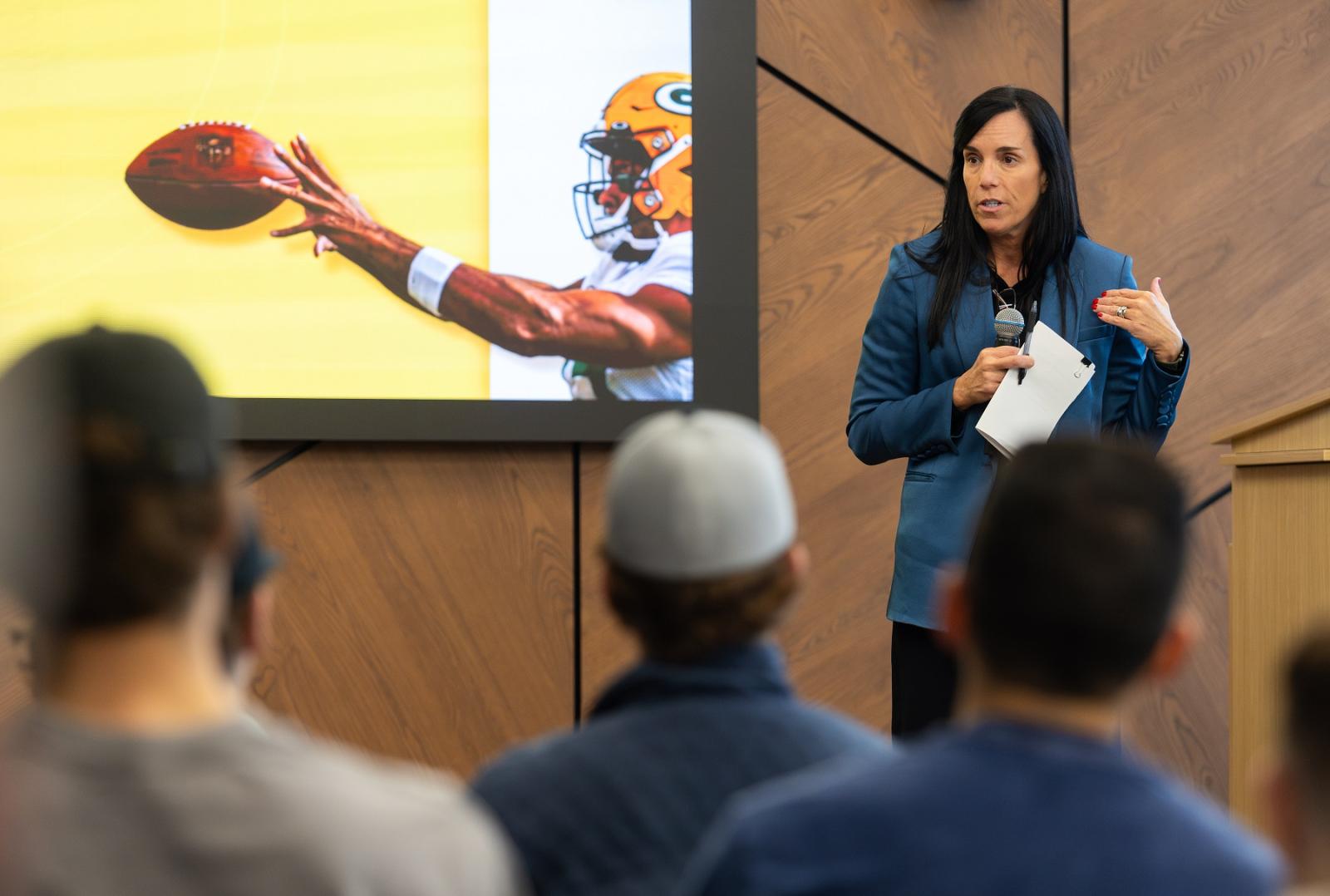 Gabrielle Valdez Dow speaks to the class of Sport Management at the Business and Entrepreneurship Center in Fox Commons. An image behind her shows a Packers player.