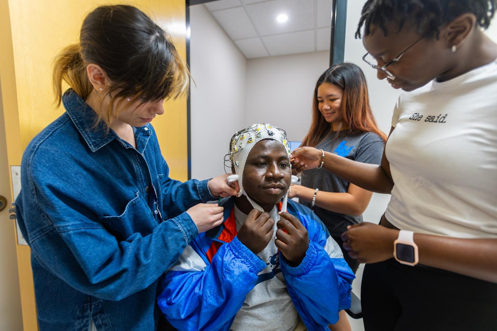 Vivian Khan, Khayla Santiago, and Fanta Jatta place an EEG cap on Den Boakye as part of summer neuroscience research.