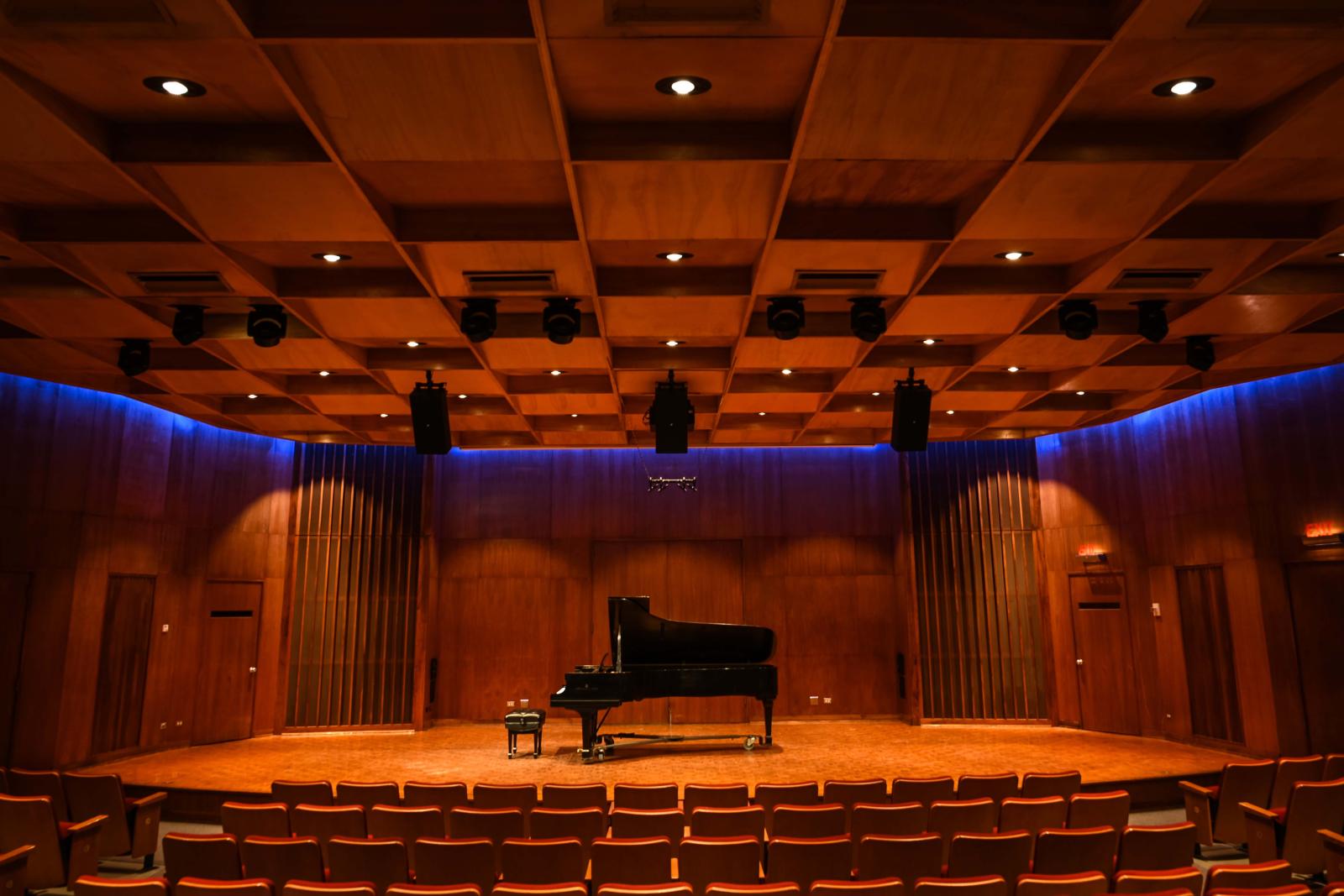 A grand piano and bench on the stage of a recital hall with wood walls, stage, and ceiling, with a hint of blue lighting tracing the seam between wall and ceiling