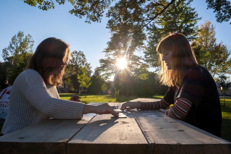 Two students sit at picnic table playing cards as sun shines through trees on campus.