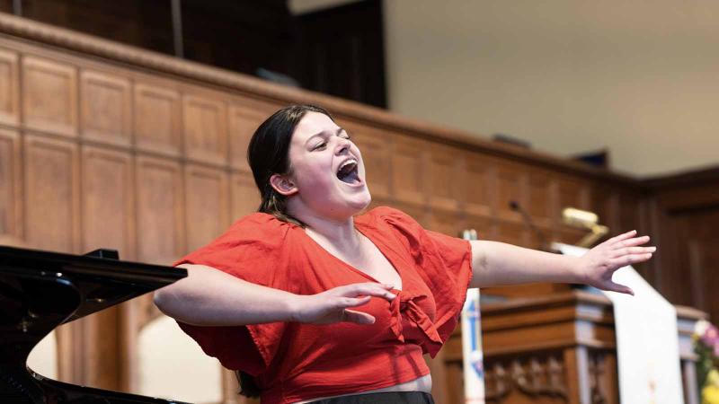 A dark-haired woman in a red shirt sings and gestures expressively