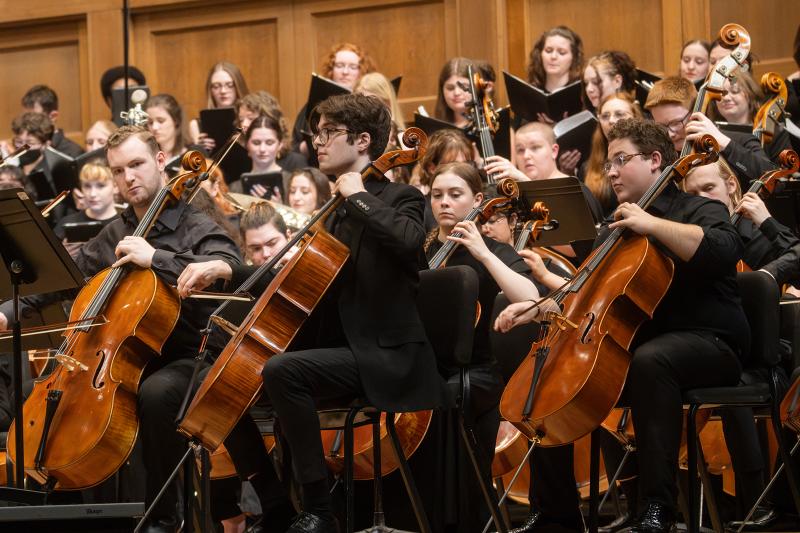 A group of musicians playing cello, with choral singers in the background.