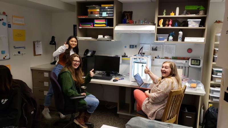 Three students in the makerspace smiling and giving a thumbs-up while sitting or standing near one of the 3D printers.