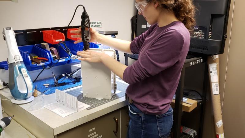 Woman standing at a workspace in the makerspace using a Dremel sander to smooth a hole in an object she 3D printed.