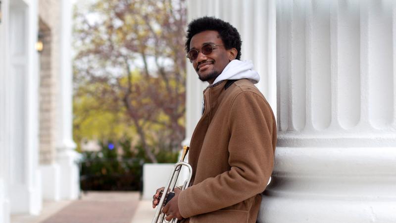 Earl Simons leans against a white column, holding a trumpet and smiling at the camera.