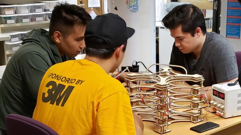 Three students sitting around a table in the makerspace looking at a wooden laser harp they constructed.