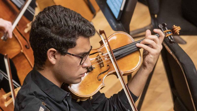 A young man in a black shirt with short dark hair and glasses is seen in profile playing the violin during an orchestra concert