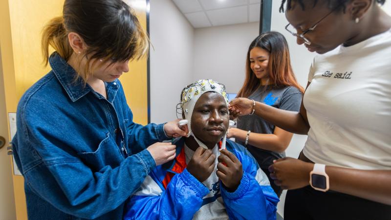 Vivian Khan, Khayla Santiago, and Fanta Jatta place an EEG cap on Den Boakye as part of summer neuroscience research.
