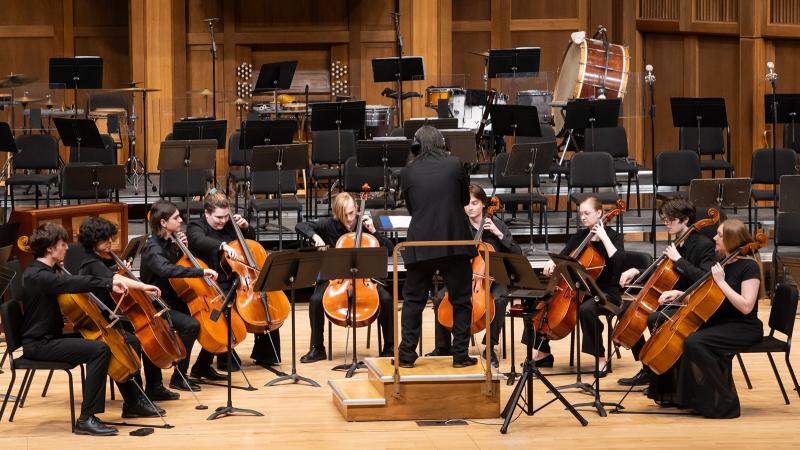 A cello ensemble performing on stage, led by a conductor.