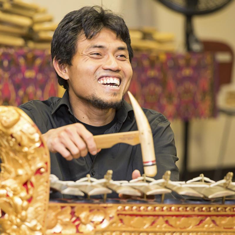 Smiling Balinese man with a short goatee playing a metallophone with a wooden mallet.