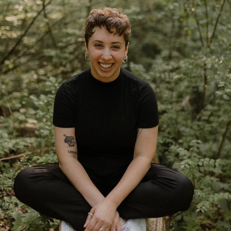 A picture of Dr. Dyer wearing a black shirt, black pants, and white sneakers. She is sitting cross-legged on a long. Behind her are green trees. She is smiling and looking at the camera.