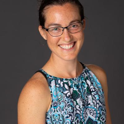 Joyful bespectacled and freckled professor with a broad smile, sitting for a portrait in a blue paisley blouse.