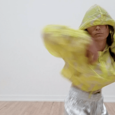 mauriah is in a yellow hooded windbreaker and silver pants. her right arm is curved inwards towards chest, hood is up covering eyes, body a bit blurred as if caught mid turn